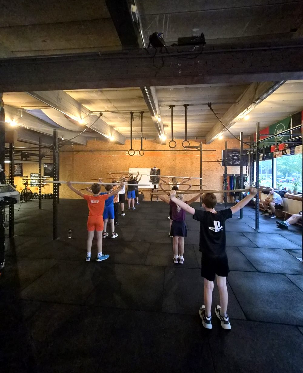Kinderen van CrossFit Teens trainen met een stok boven het hoofd op de vloer van de box, gezien van achteren